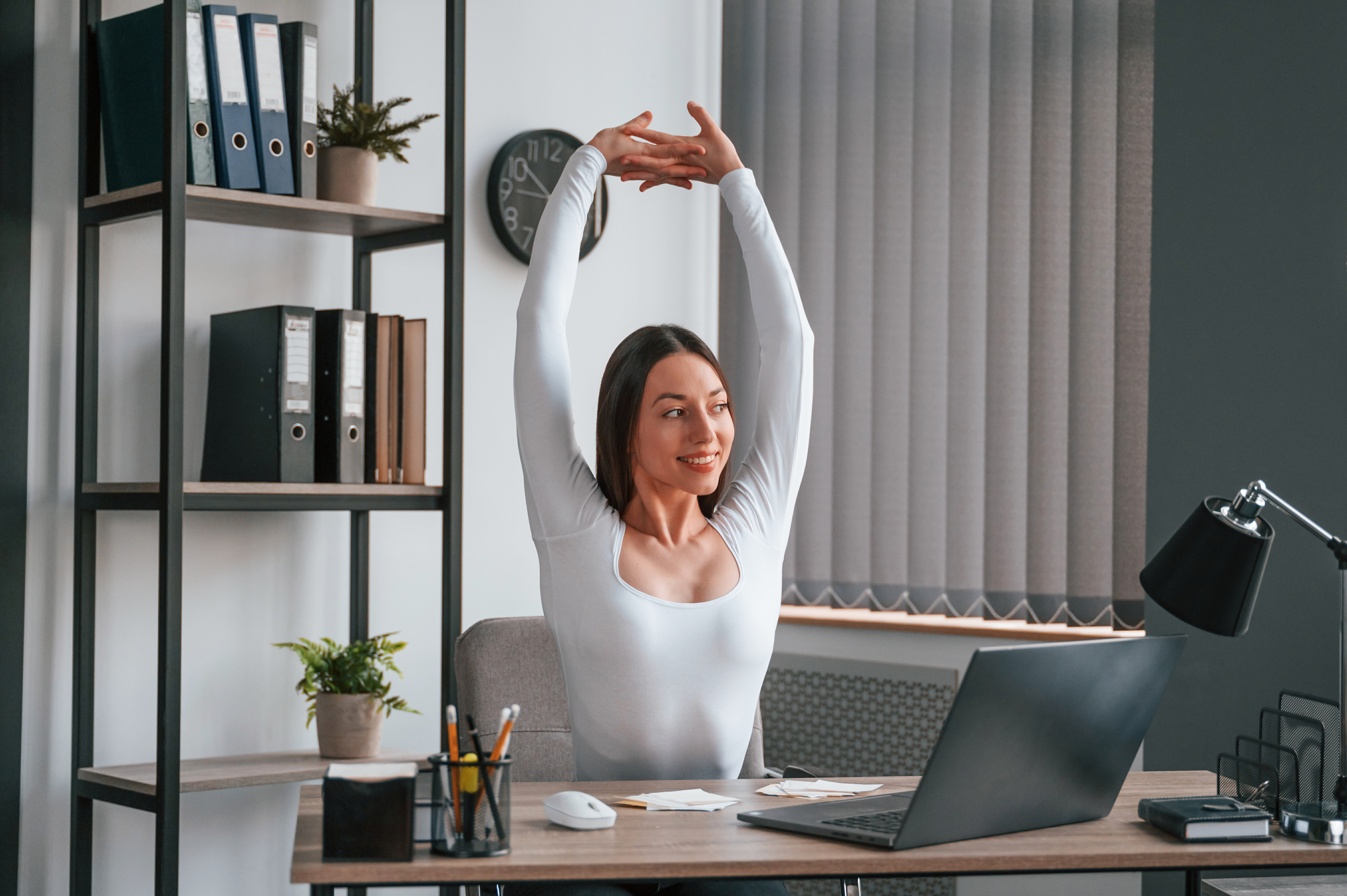 Una mujer profesional estirando los brazos detrás de su escritorio en el trabajo, tomando un descanso para mejorar la postura y aliviar la tensión.