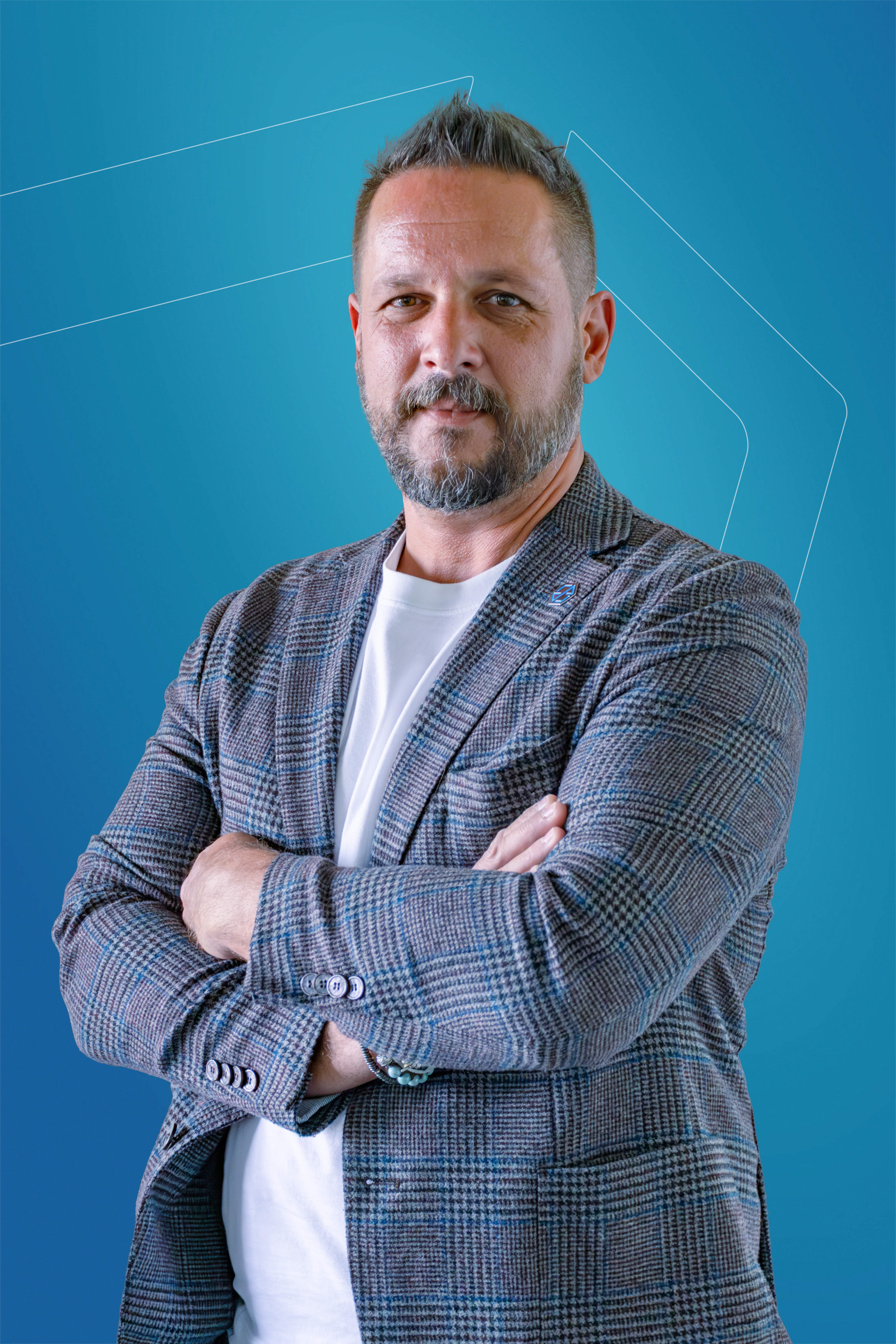 Professional portrait of a man in a checked blazer with arms crossed, standing against a blue gradient background, photographed for Timeless Tech corporate materials.